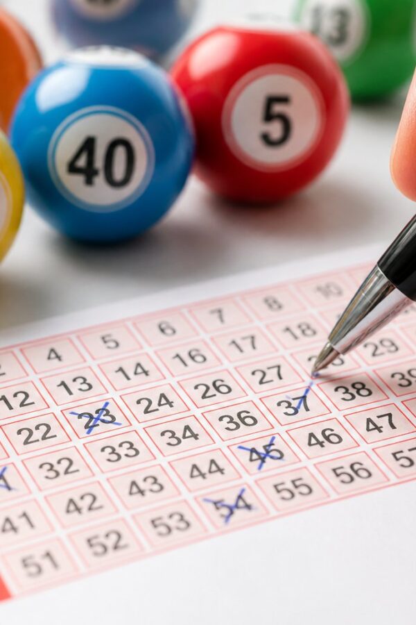 Mão marcando números em um bilhete de loteria cercado por bolas numeradas sobre uma mesa.
